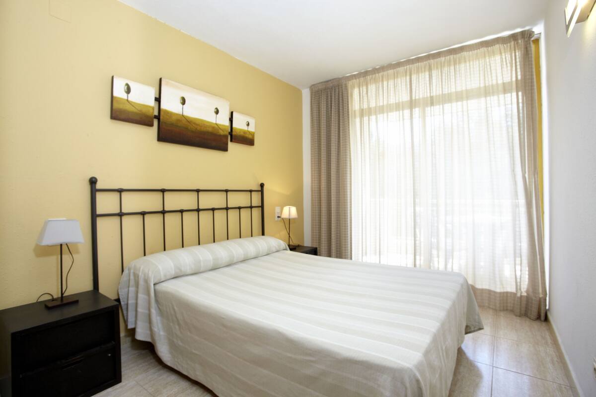 A hotel room with a single bed, a nightstand with a lamp, and a window with sheer curtains.