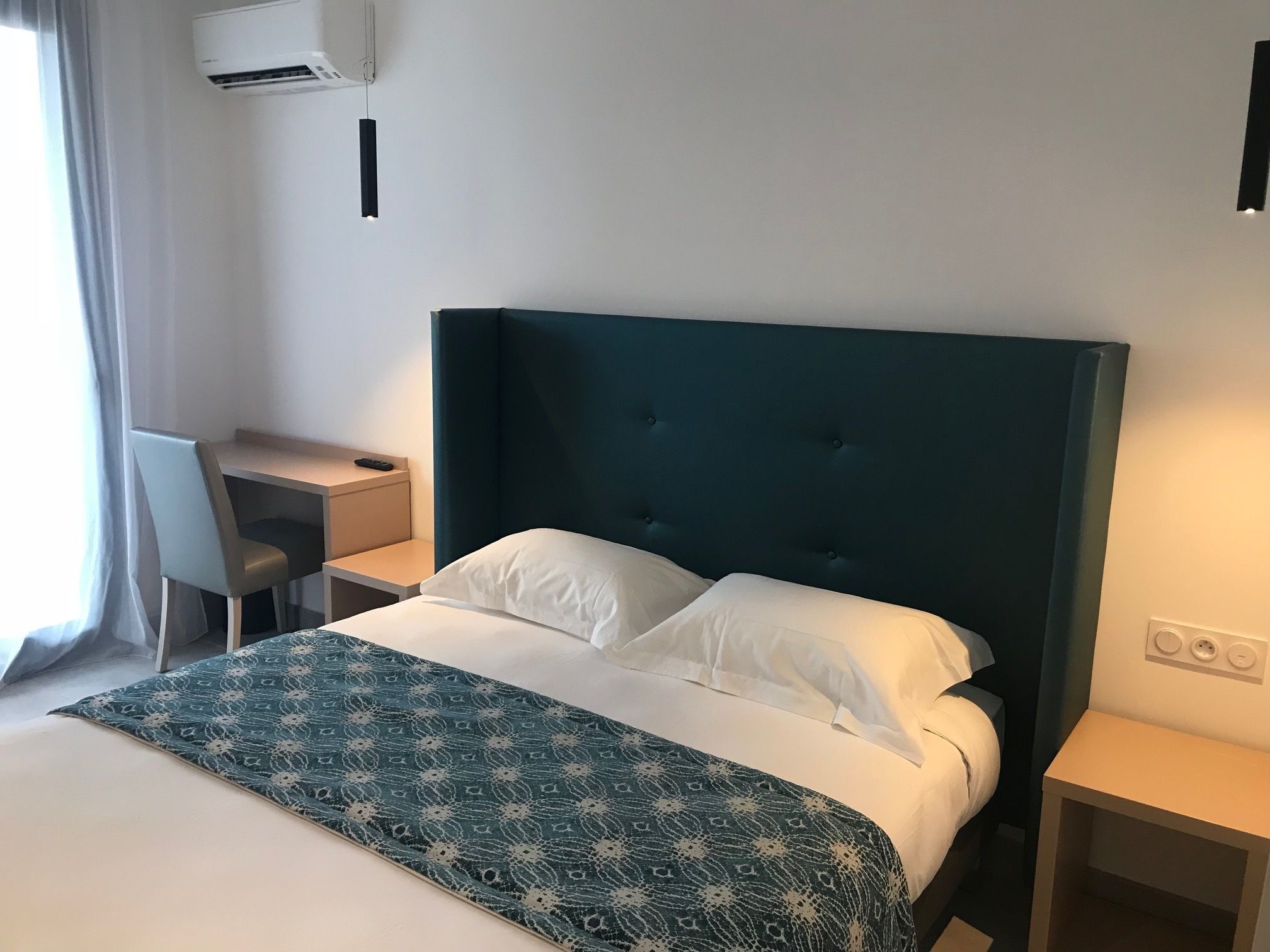 A hotel room with a bed, a desk, a chair, and an air conditioning unit.