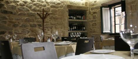 A dining area with stone walls, tables set with white tablecloths, and wine bottles on shelves.