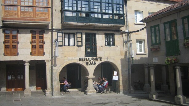Un edificio con un letrero que dice "RESTAURANTE RUAS" y dos personas sentadas afuera.