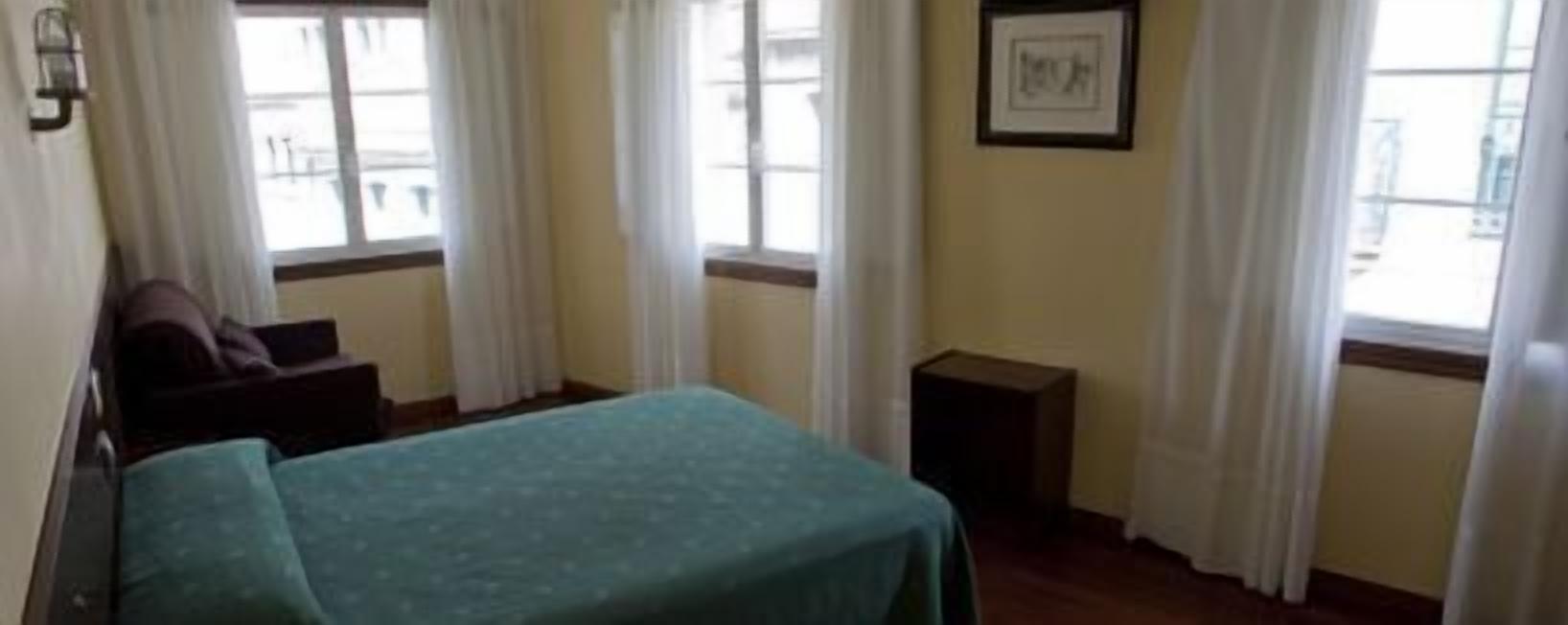 Una habitación con una cama, una silla, una mesita y una ventana con cortinas.