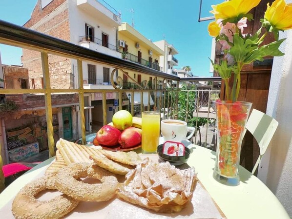 A table with breakfast items including apples, a glass of orange juice, a cup of coffee, and pastries on a balcony with a view of buildings and a clear sky.