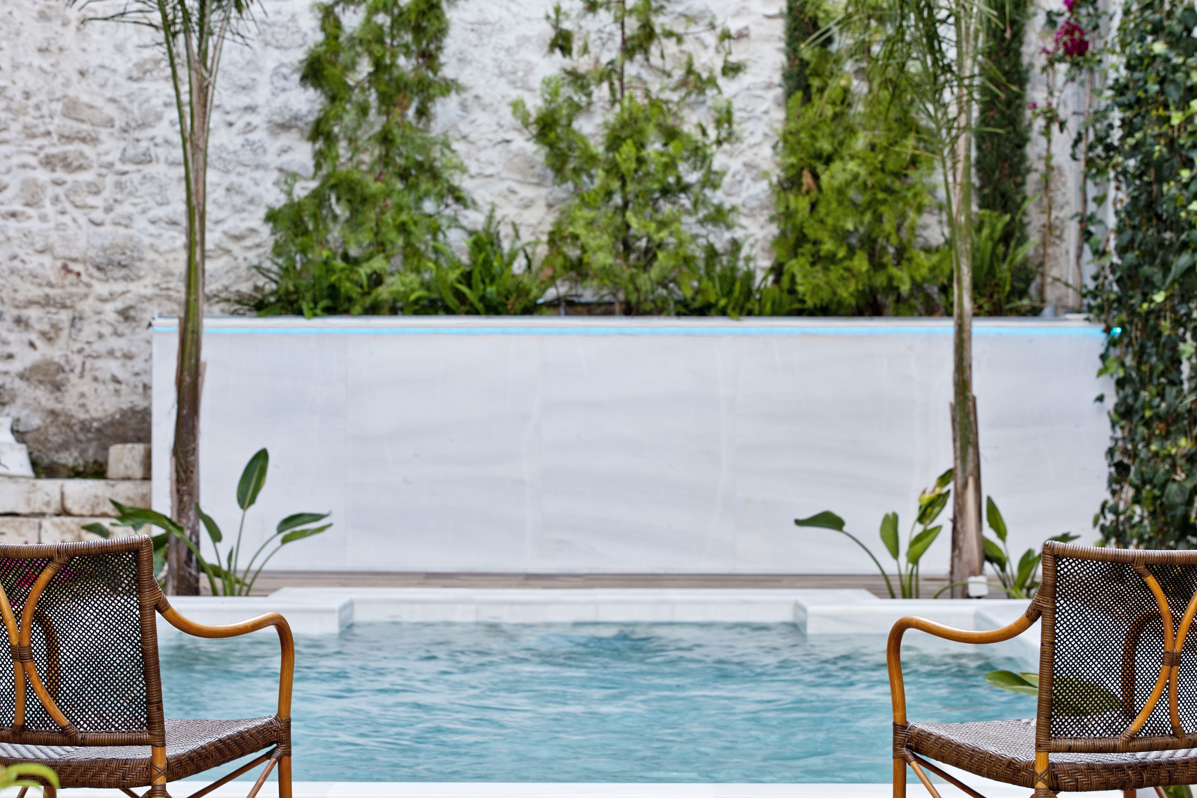 A pool area with two wicker chairs facing the water, surrounded by greenery and a stone wall.