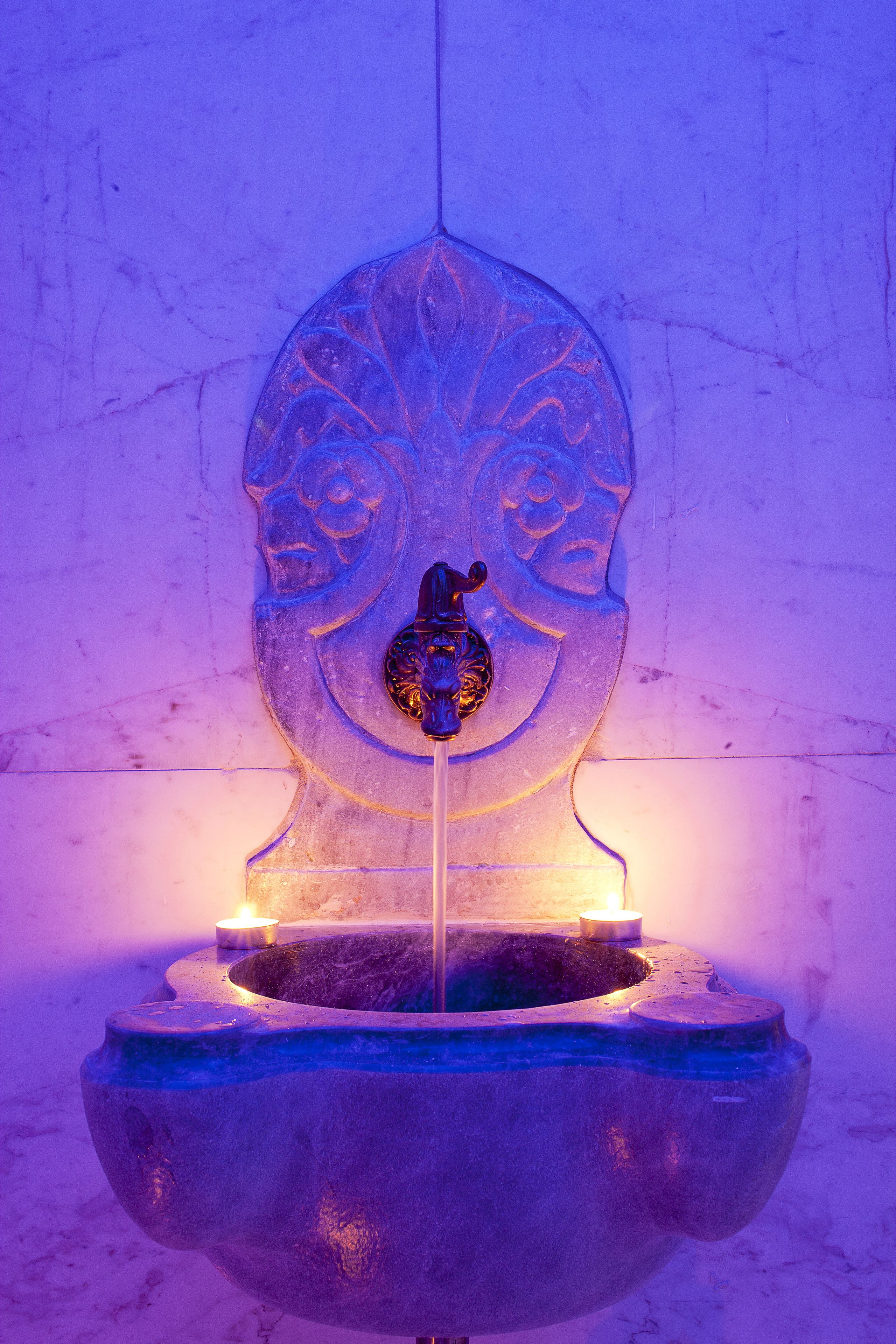 A stone fountain with a central spout and a decorative headpiece, illuminated by a candle.