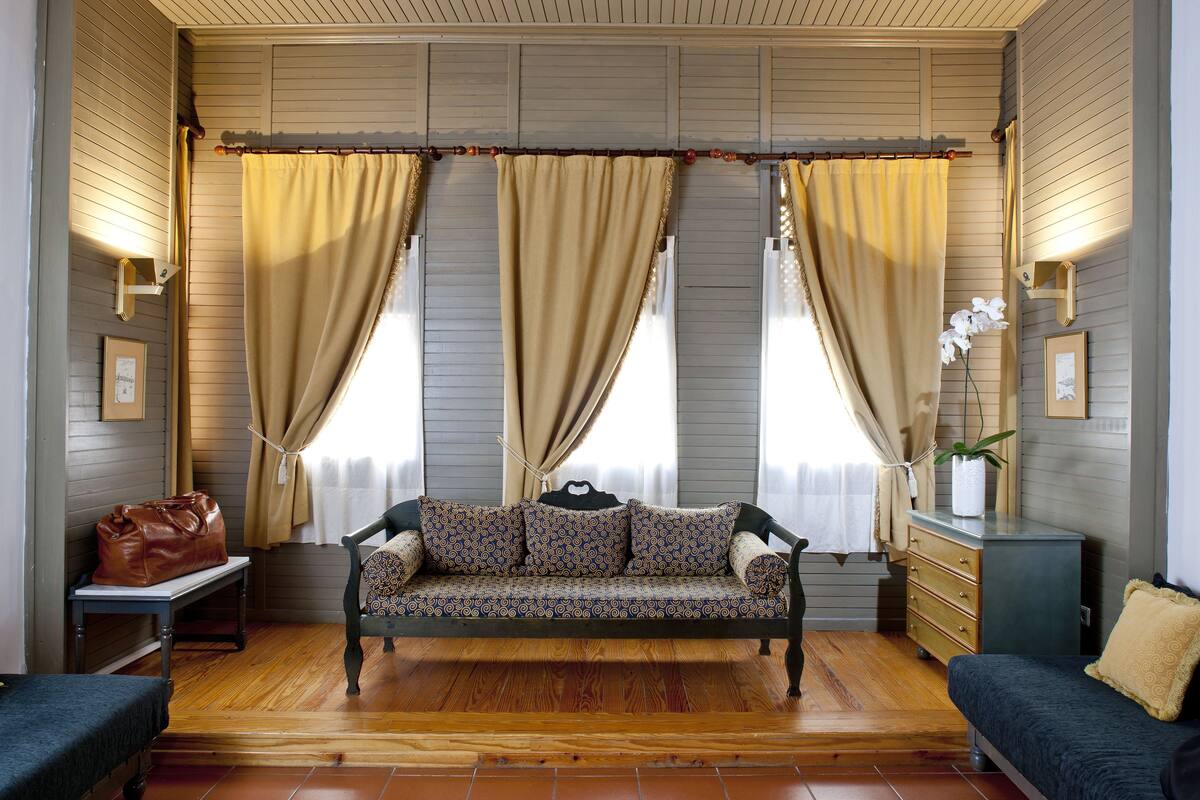 A room with a wooden floor, a sofa with patterned cushions, a bench, a dresser, and a bench with a bag on it.