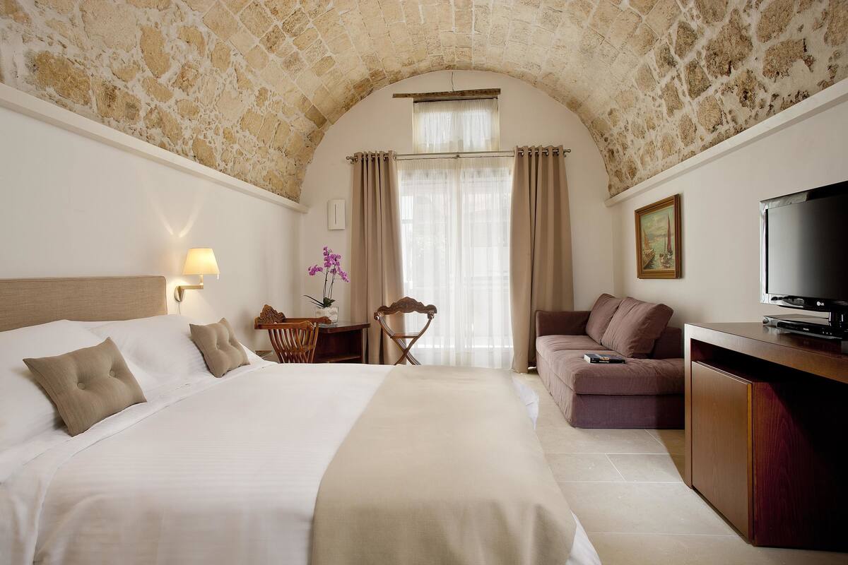 A hotel room with a large bed, a desk, a chair, a sofa, a TV, and a stone ceiling.