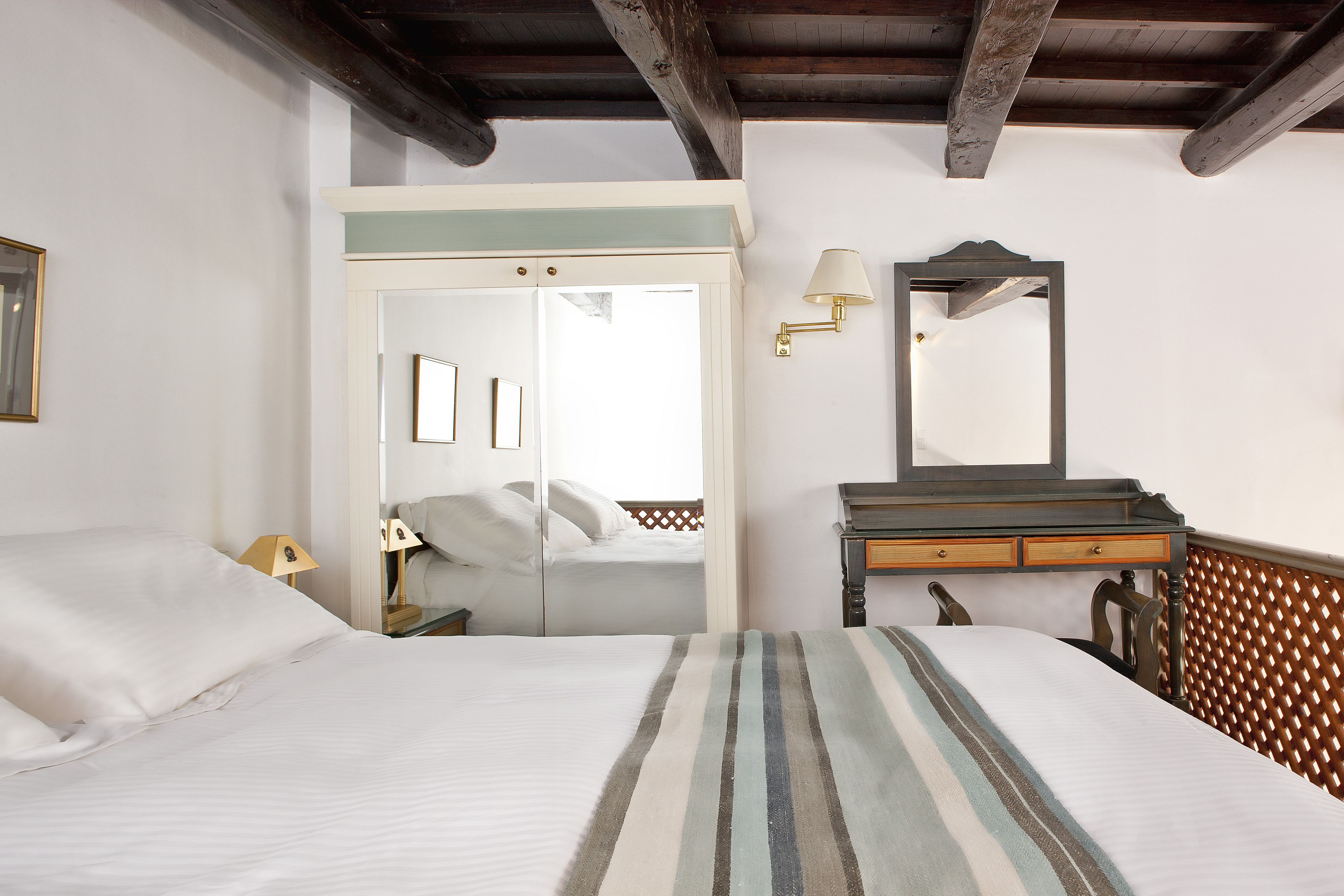 Un dormitorio con cama, mesita de noche, cómoda, espejo y una puerta que da a otra habitación.