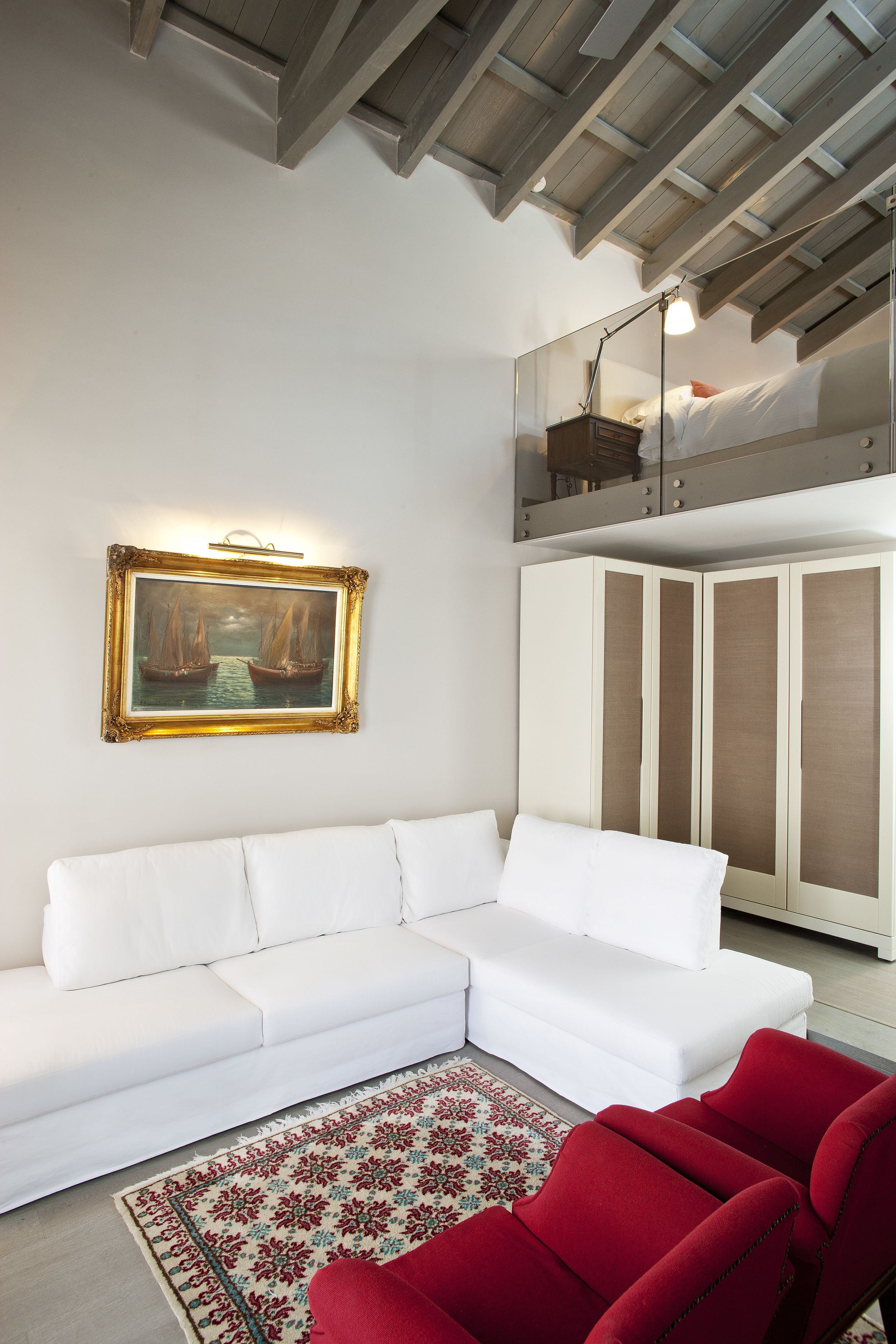 A white sectional sofa, a red armchair, a patterned rug, a framed painting, and a mirrored wardrobe.