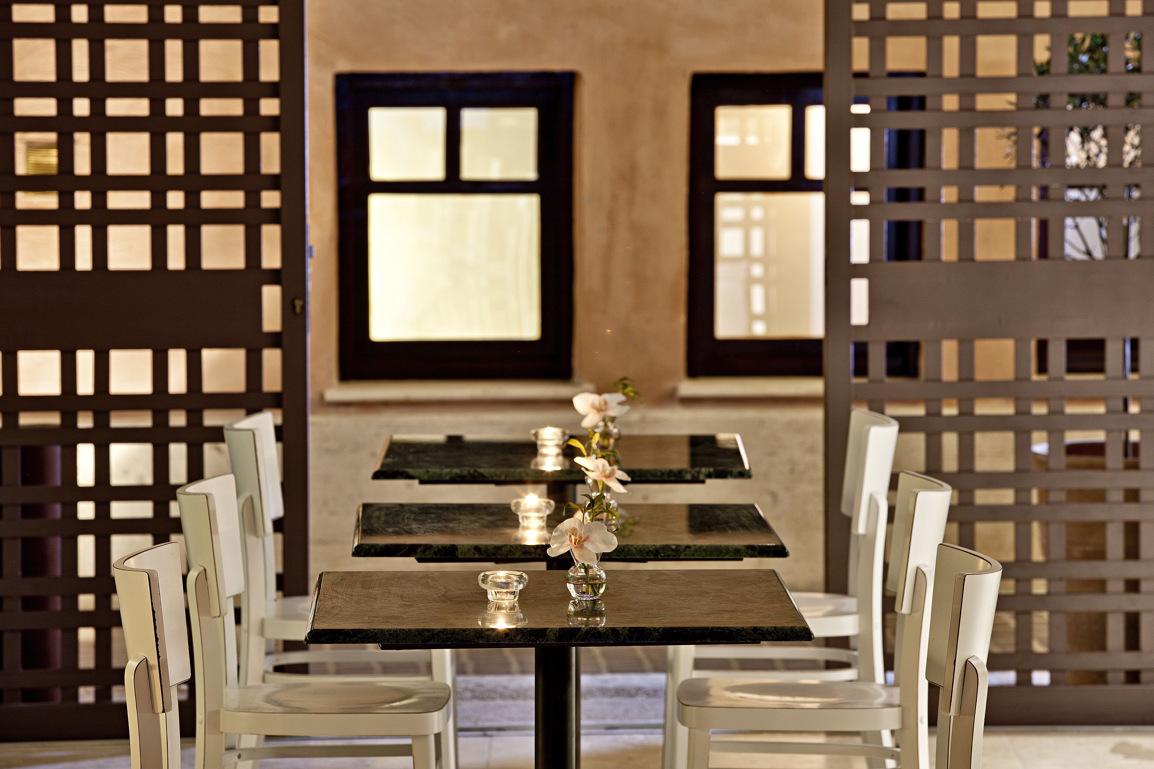 Un coin repas avec une table dressée pour quatre personnes, entourée de chaises blanches. On y voit deux fenêtres à cadres foncés, et un paravent décoratif à motifs en treillis est visible à l’arrière-plan.