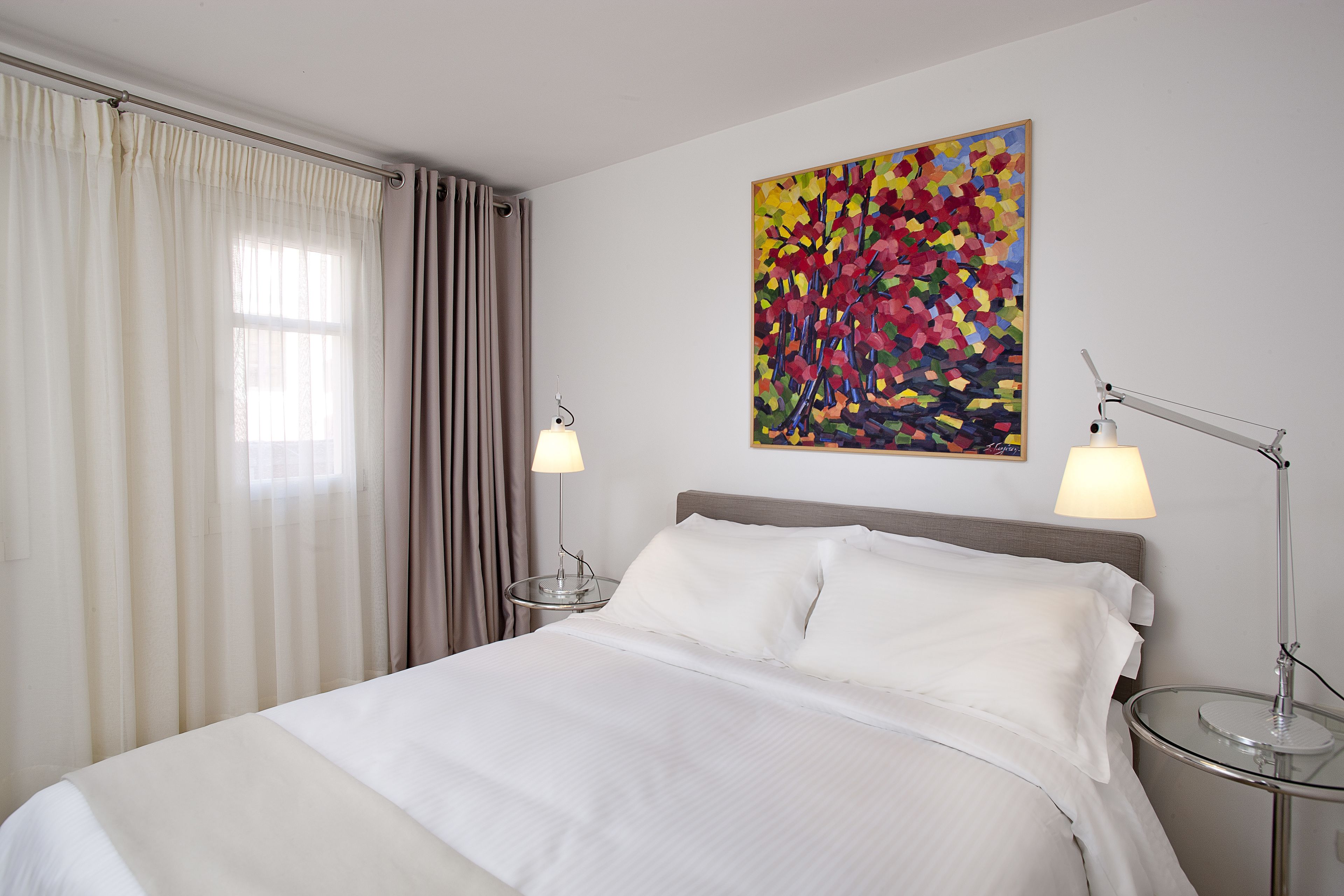 A hotel room with a bed, two bedside lamps, a painting on the wall, and a window with curtains.
