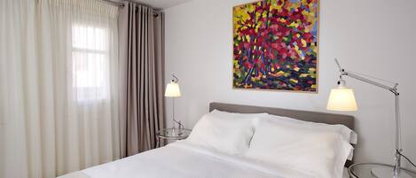 A hotel room with a bed, two bedside lamps, a painting on the wall, and a window with curtains.