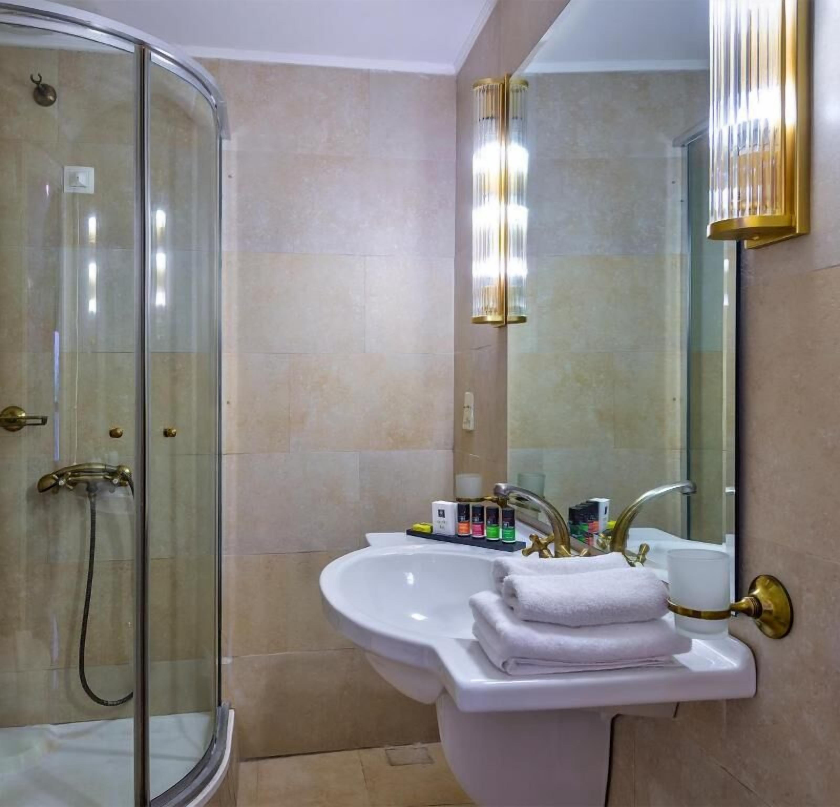 Un baño con ducha envuelta en vidrio, un lavabo blanco con detalles dorados y un espejo grande.
