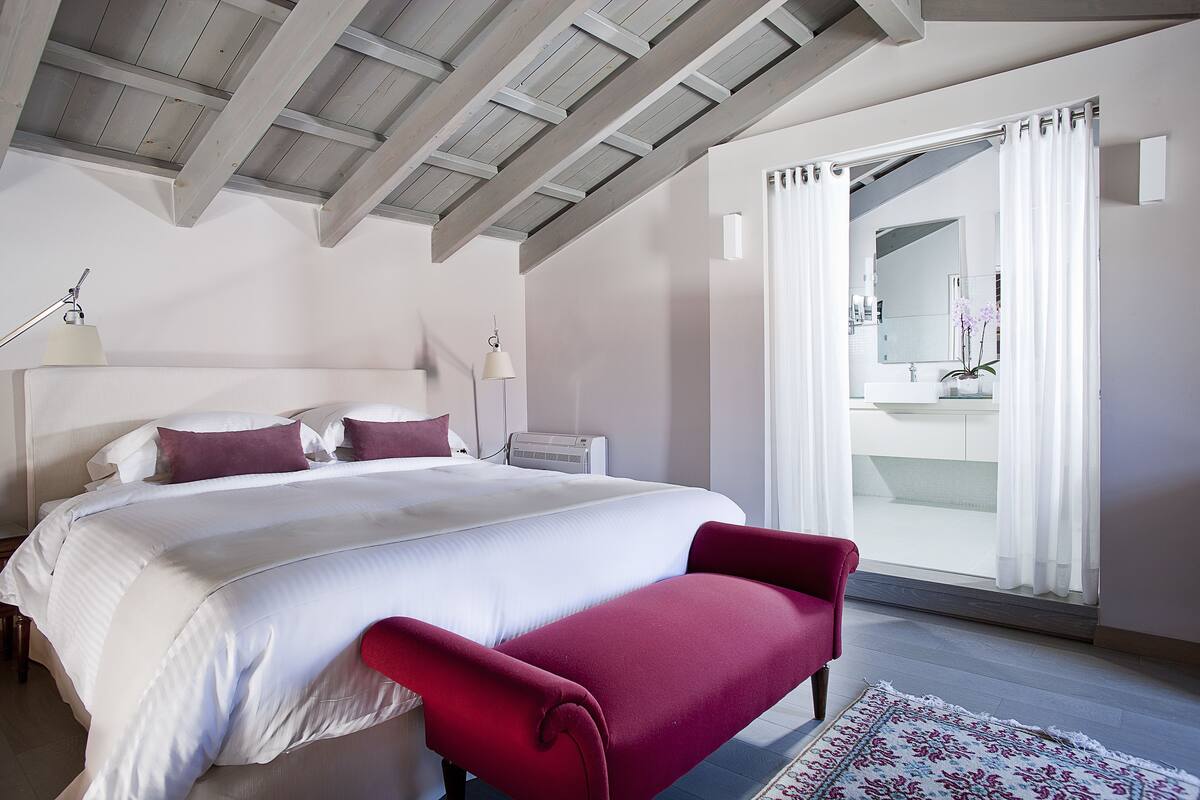 A bedroom with a large bed, a red bench, a white lamp, and a view of a bathroom through a glass door.