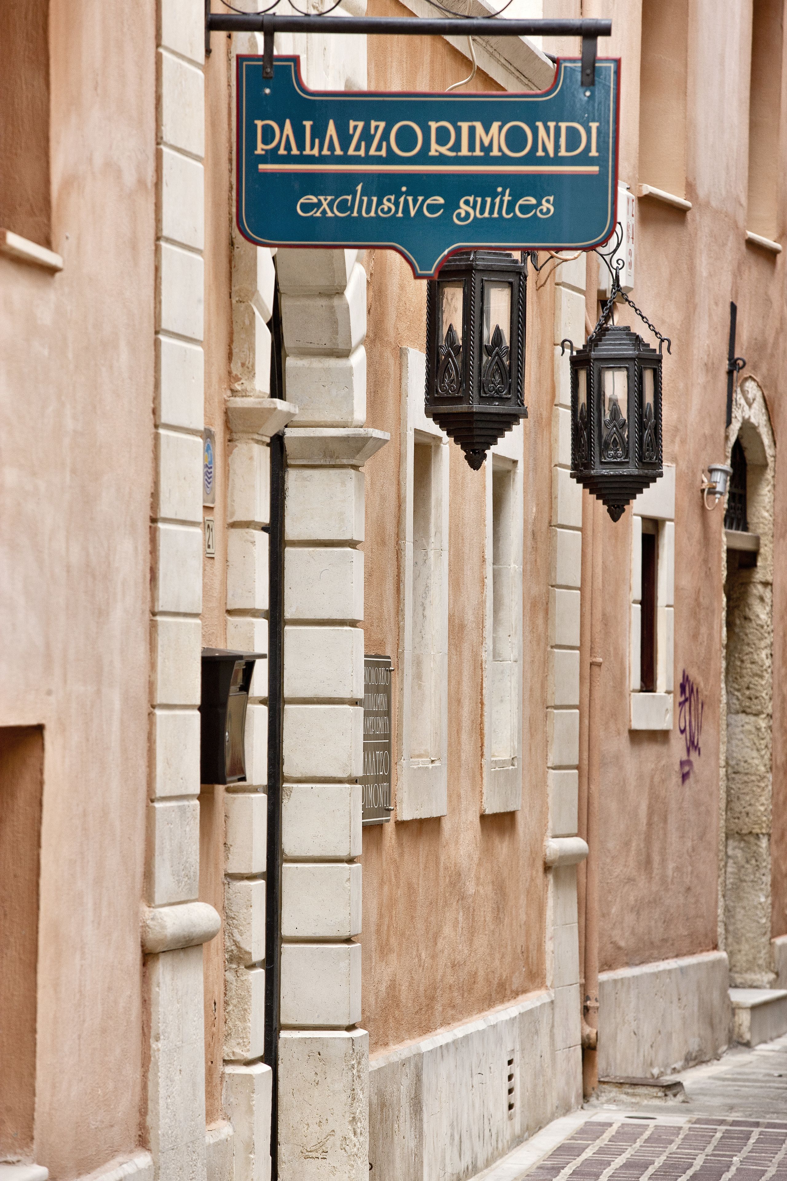 A sign for "Palazzorimondi exclusive suites" with a classic architectural style, including stone walls and decorative lanterns.