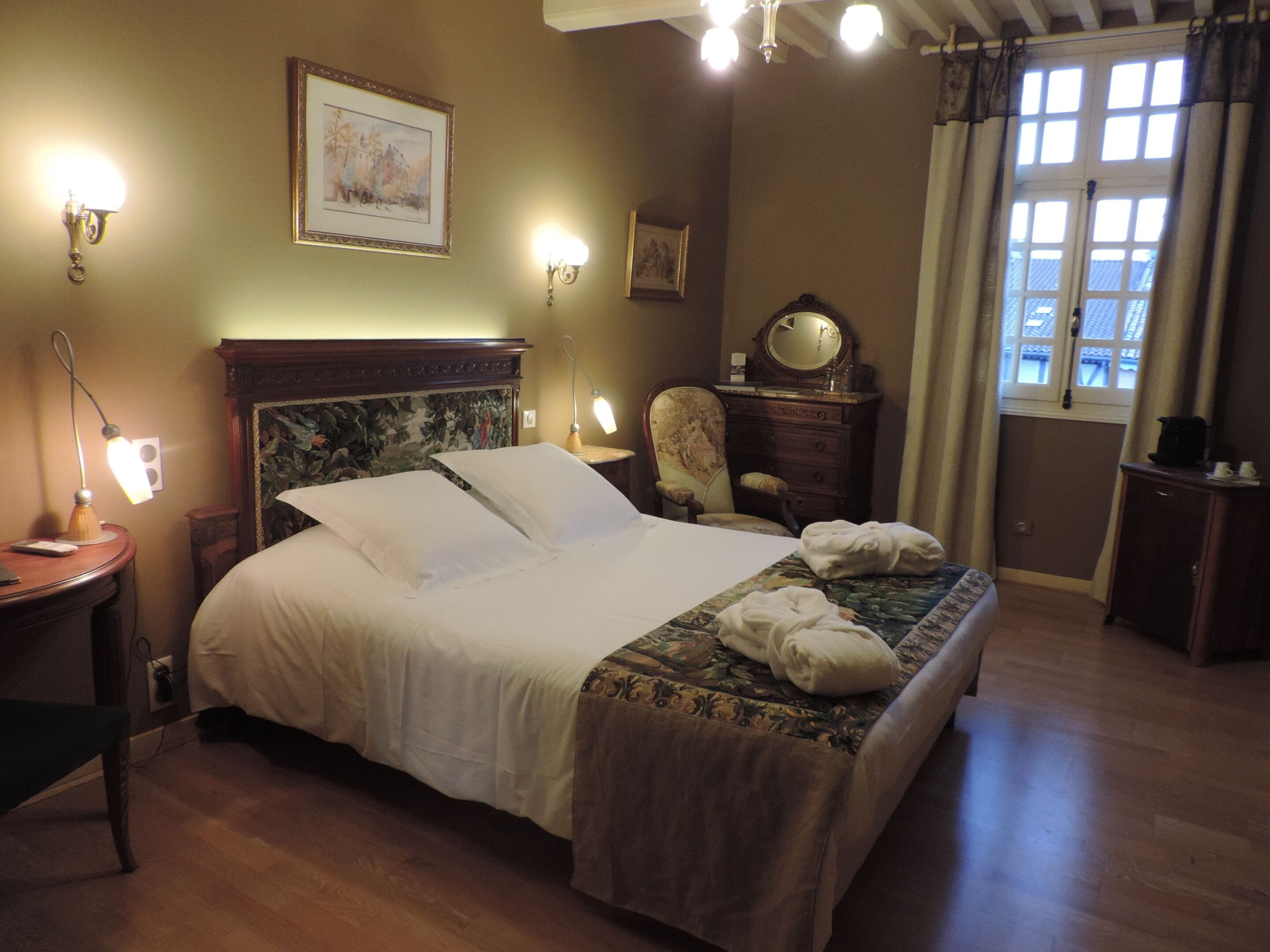 Een net opgemaakt bed met hoofdbord, een stoel, een dressoir en een raam met gordijnen.