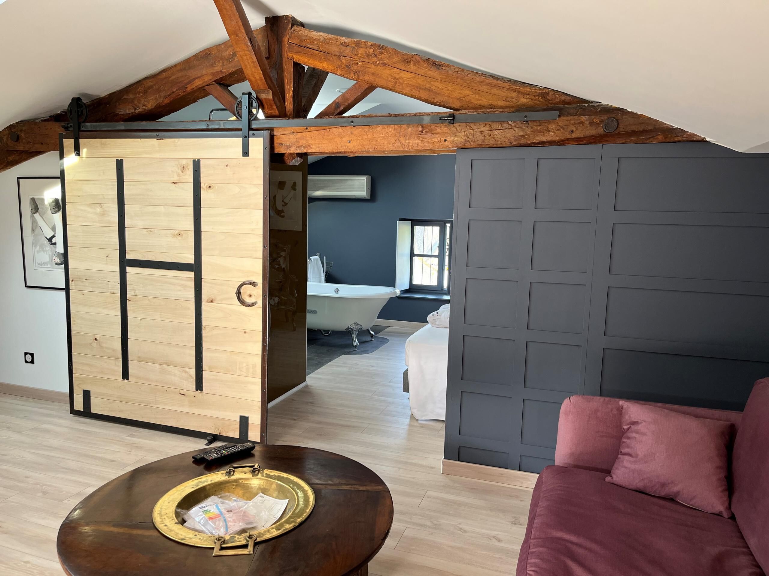 A loft-style room with exposed wooden beams, a sliding door, a round coffee table with a tray, a red sofa, and a view of a bathroom with a bathtub.