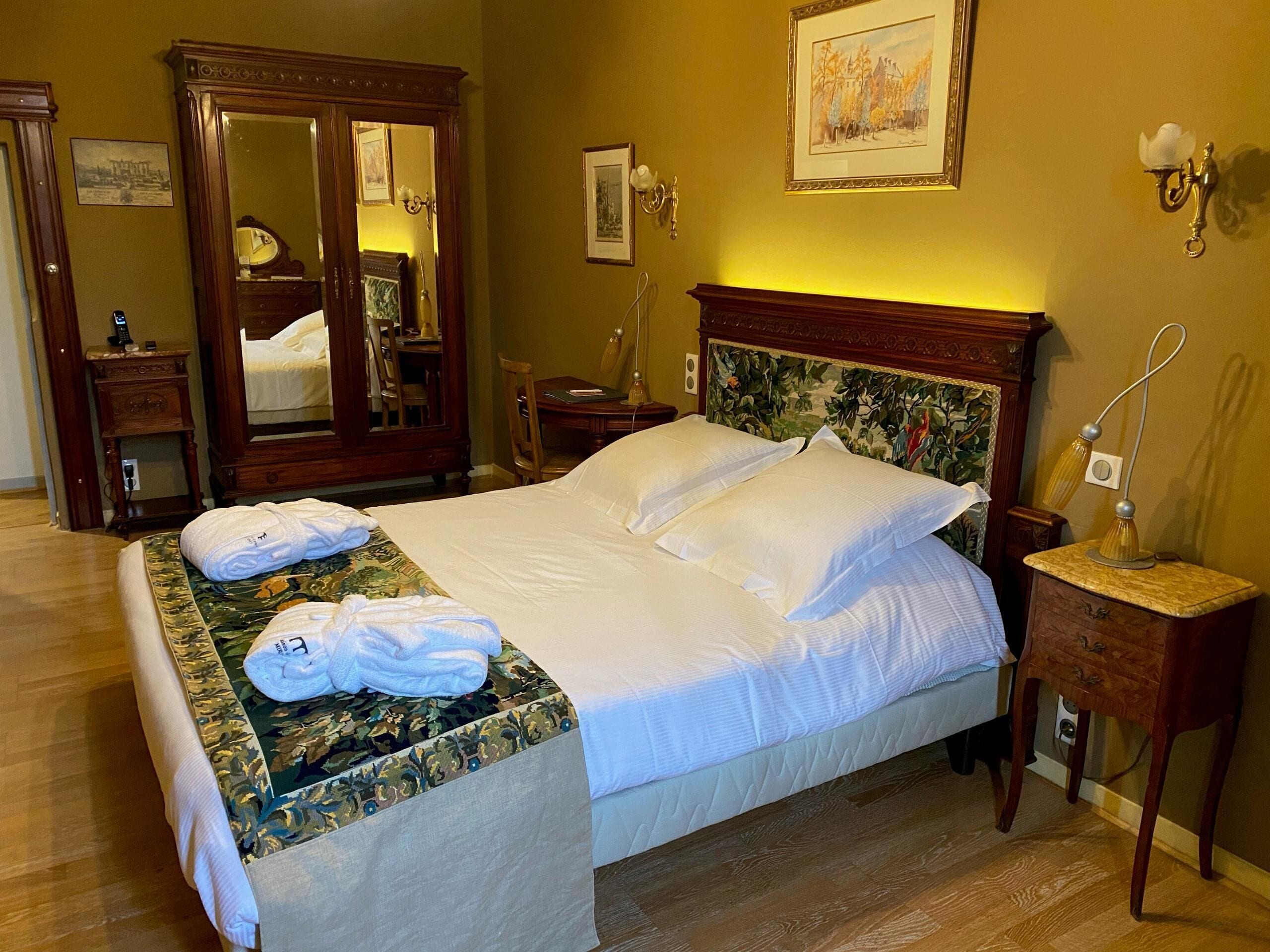 Una cama bien hecha con sábanas blancas, un cabecero de madera con motivos florales, una mesita de noche con una lámpara y un espejo sobre la cama.