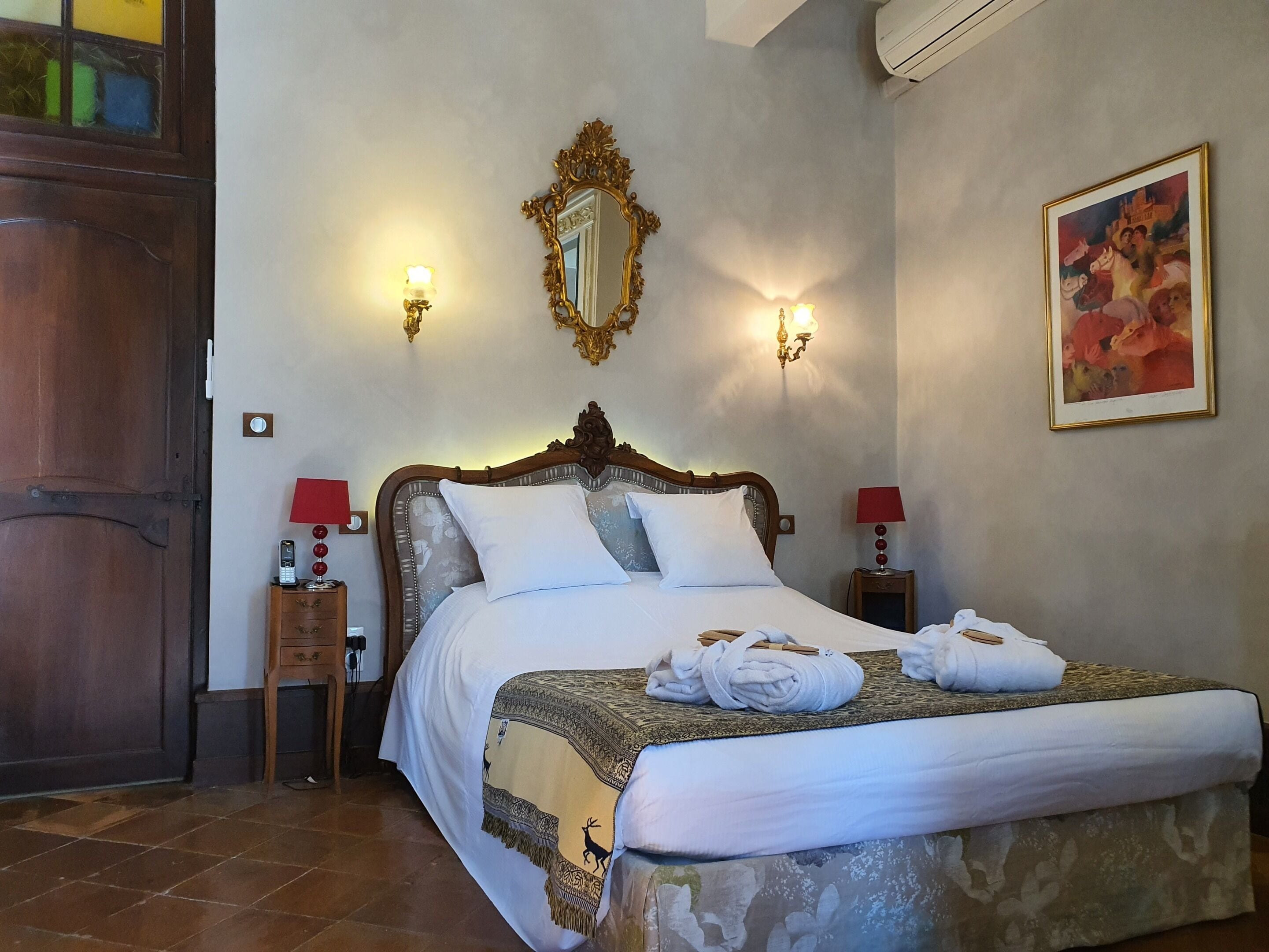 A neatly made bed with a patterned blanket, two bedside lamps, a wall-mounted mirror, and a painting.