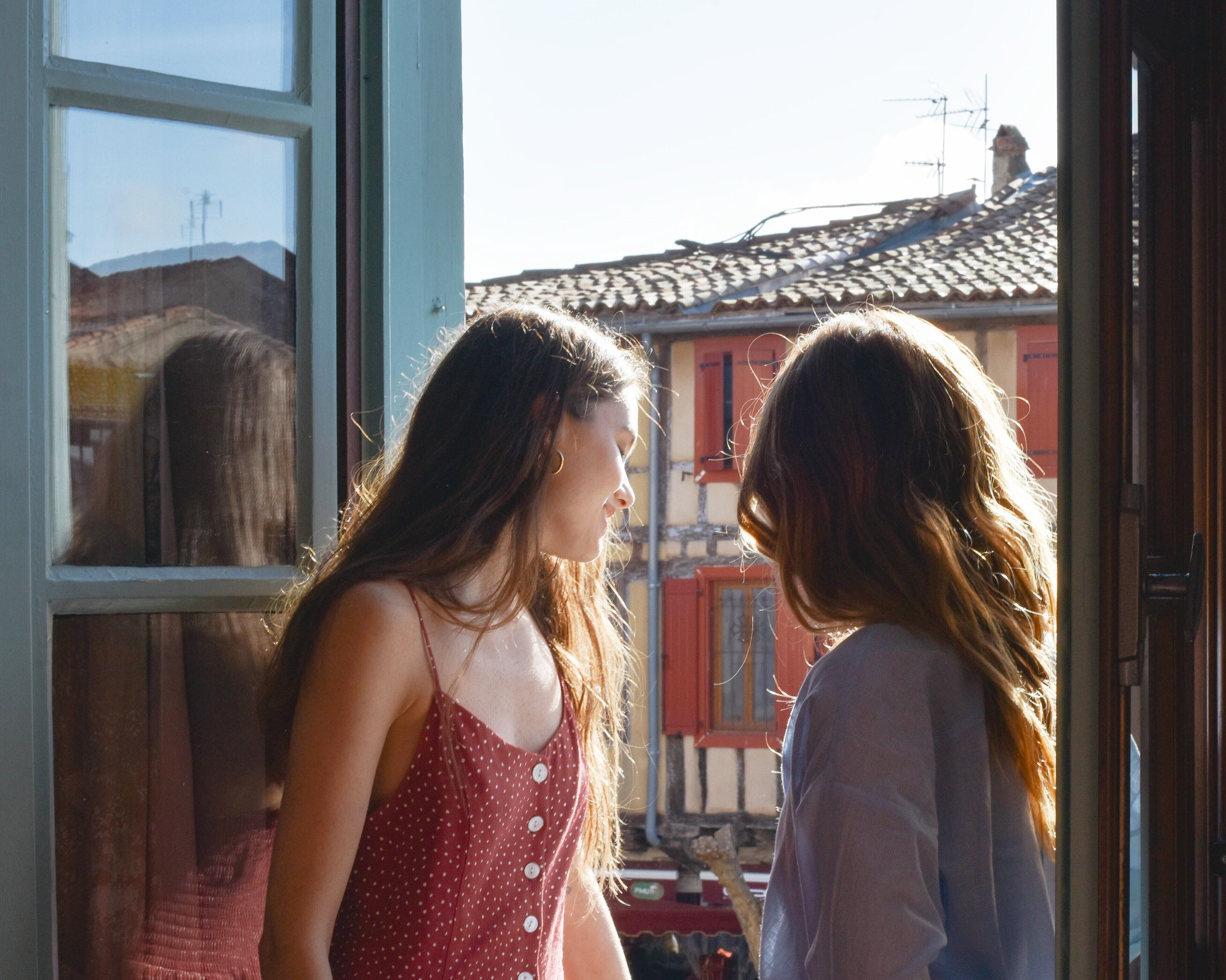 Two people standing at a window with a view of buildings and a tiled roof.