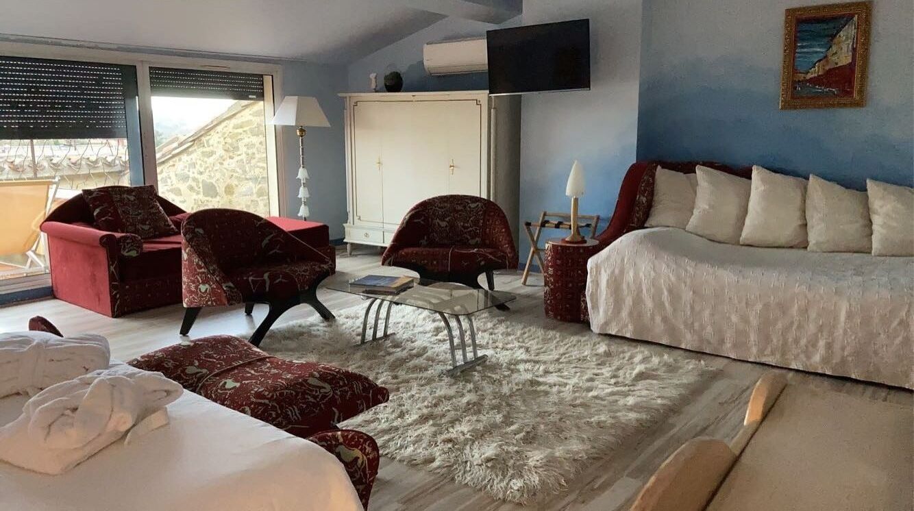 A hotel room with a bed, two red armchairs, a small table, a white sofa, a fluffy rug, and a painting on the wall.