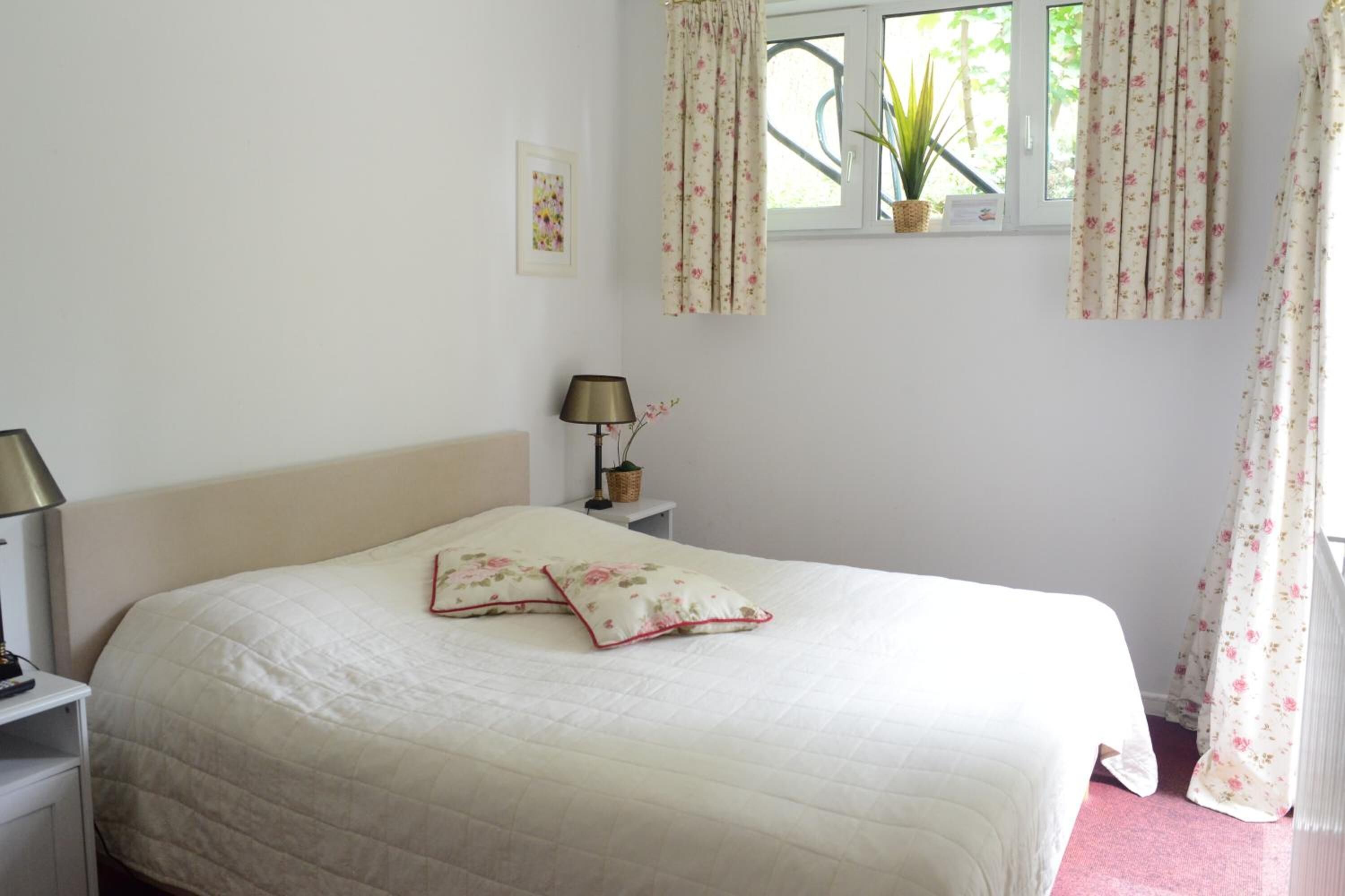 A bedroom with a bed, a nightstand, a lamp, a window with curtains, and a potted plant.