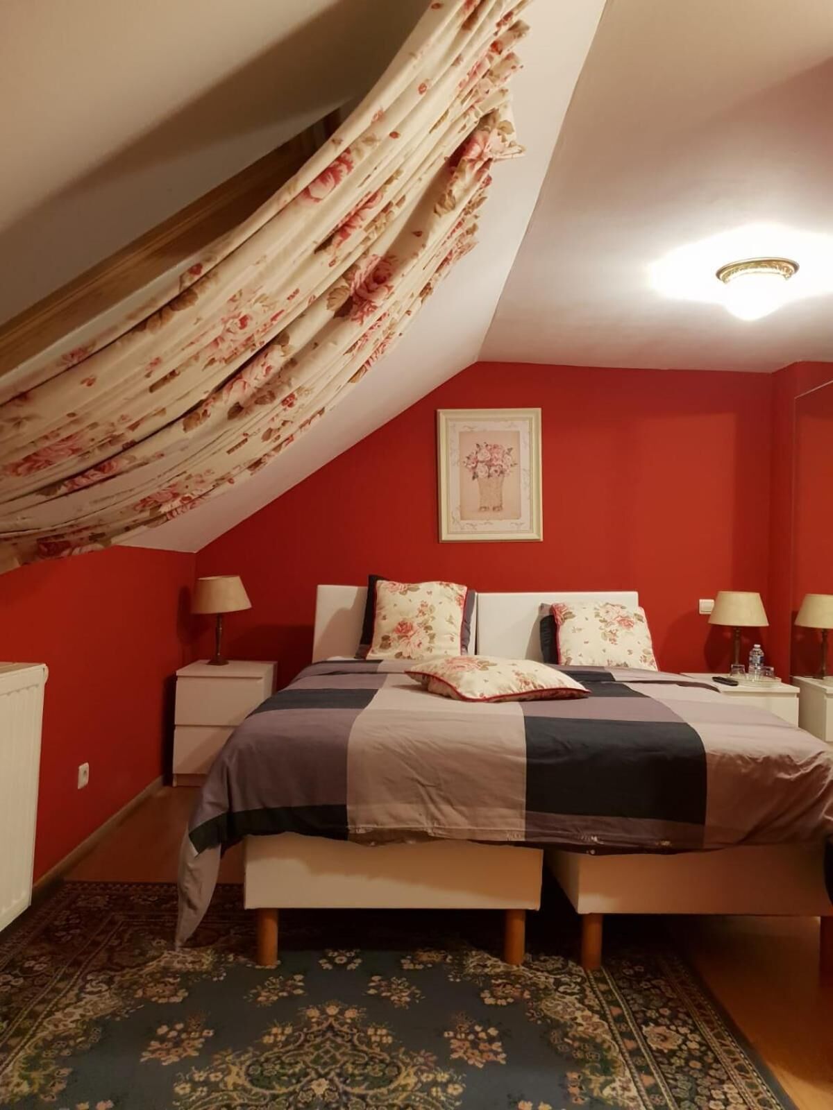 A bedroom with a sloped ceiling, red walls, a bed with floral bedding, two bedside tables with lamps, and a framed picture on the wall.
