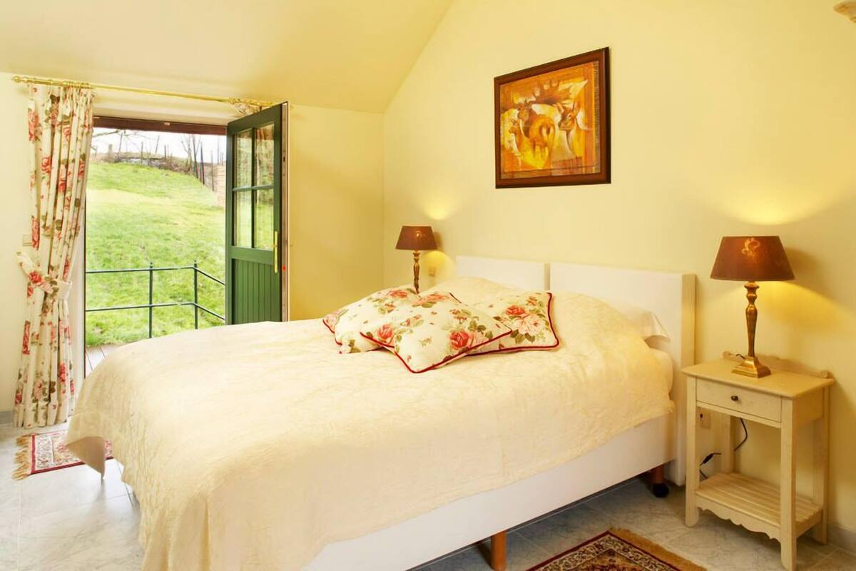 Een slaapkamer met een bed, twee bedlampen, een nachtkastje, een schilderij en uitzicht op een groen landschap door een geopende deur.