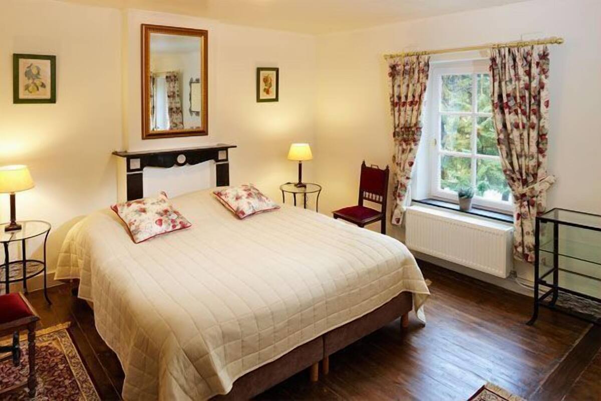 Een slaapkamer met een groot bed, twee bedlampen, een stoel, een raam met gordijnen en een radiator.