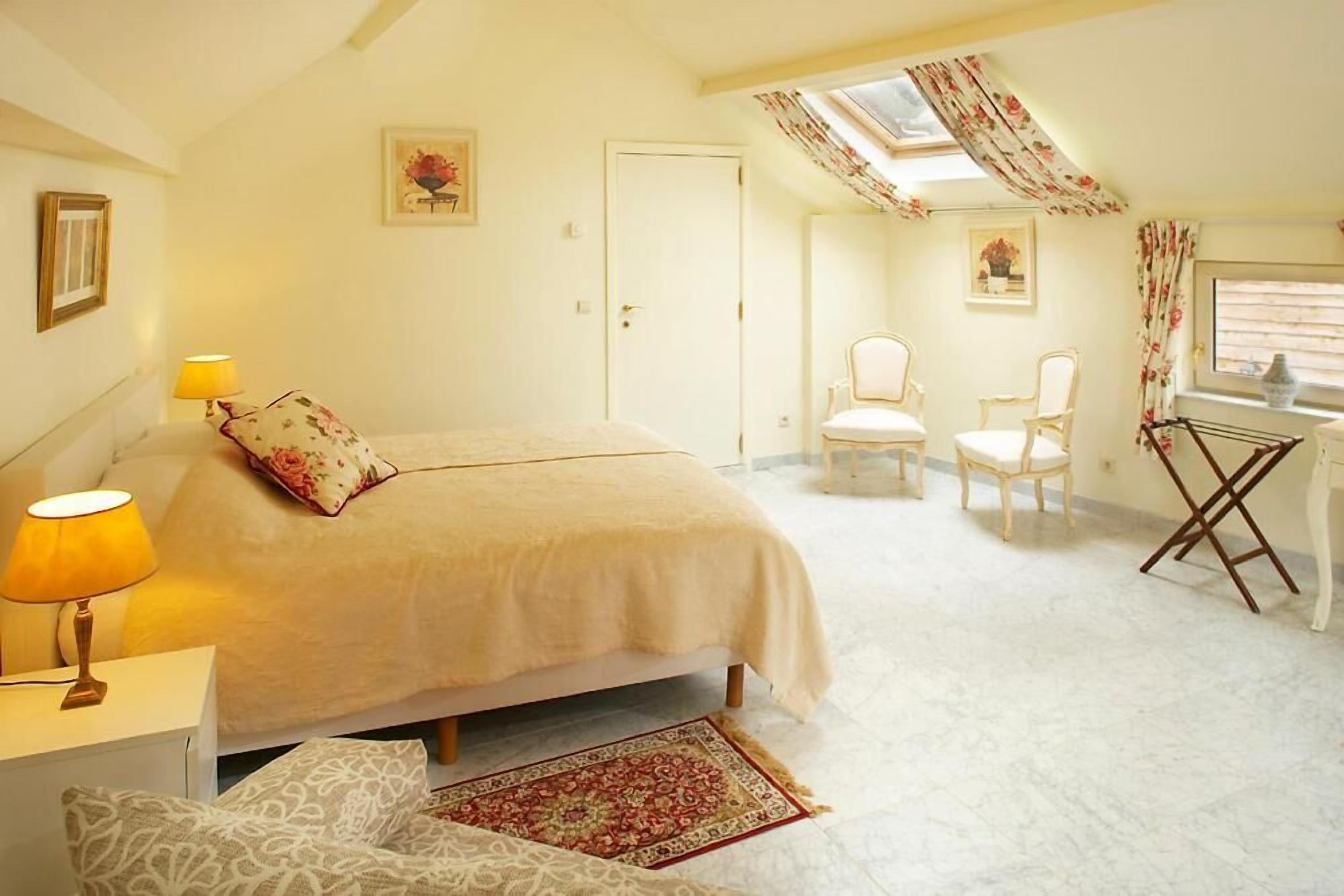 Une chambre à coucher avec un lit, deux chaises, un bureau et une fenêtre avec des rideaux.