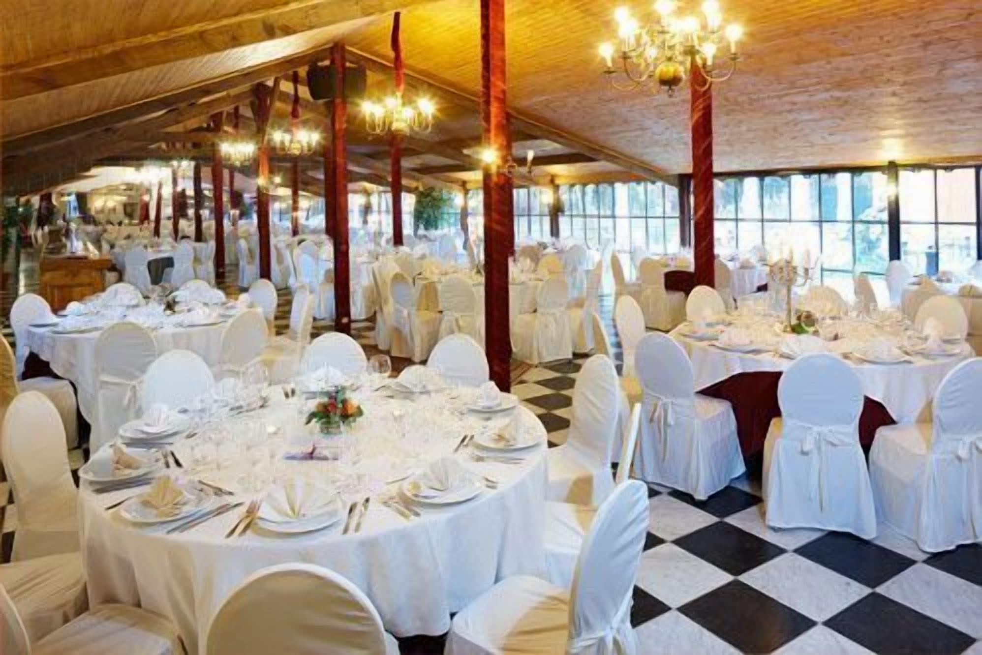 Een balzaal met ronde tafels gedekt voor een diner, witte stoelen, kroonluchters en grote ramen.