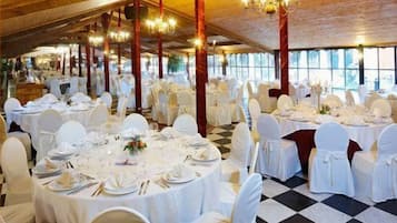 Een balzaal met ronde tafels gedekt voor een diner, witte stoelen, kroonluchters en grote ramen.