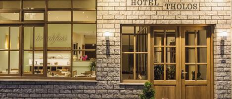 A hotel with a stone facade, large windows, and a sign reading "HOTEL THOLOS".