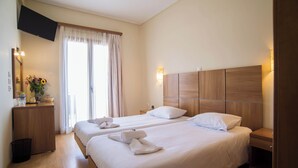 A hotel room with two beds, a wooden headboard, a TV mounted on the wall, and a window with curtains. - Tholos (Delphi)