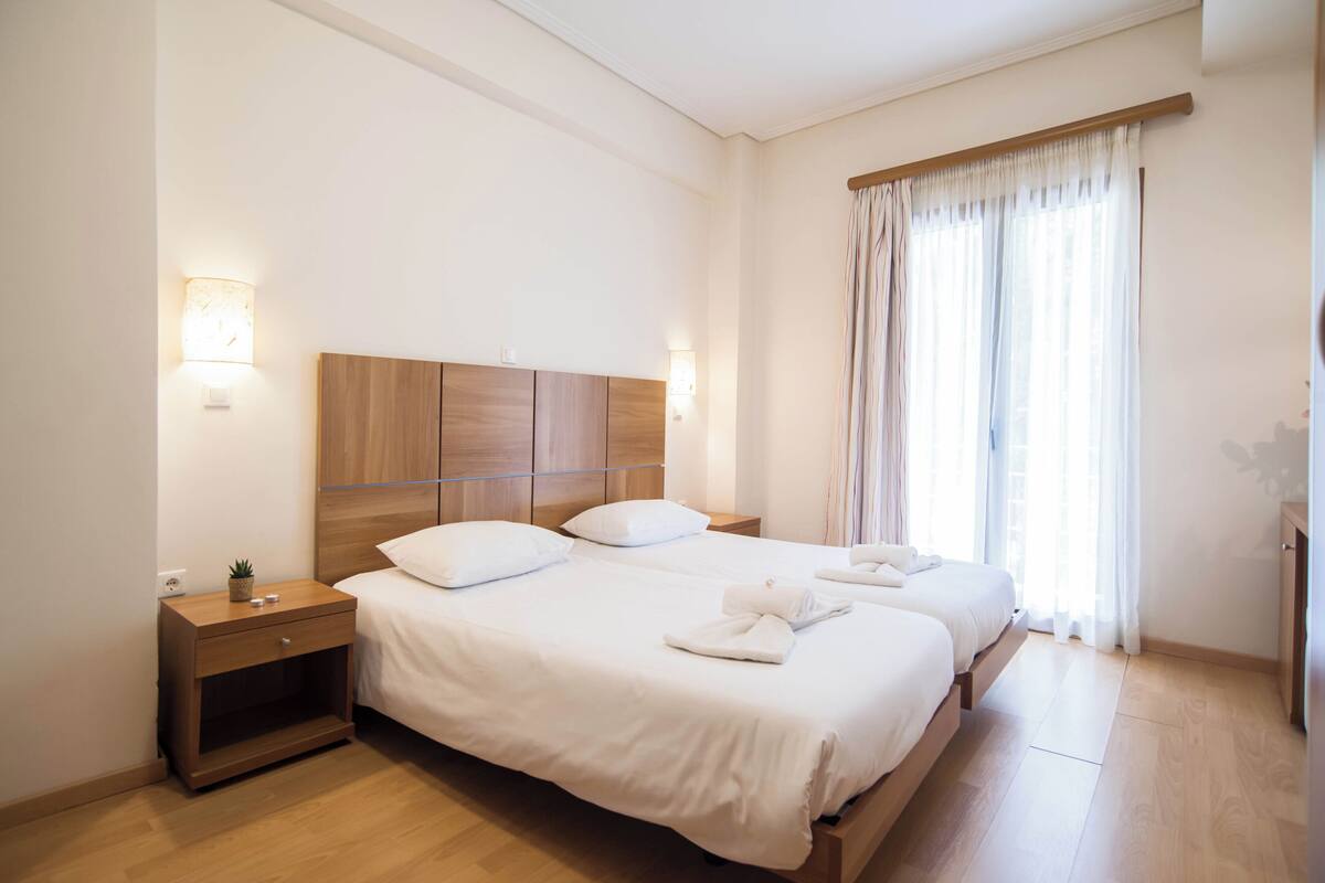 Une chambre d’hôtel avec deux lits, des têtes de lit en bois, des tables de chevet et une grande fenêtre avec des rideaux.