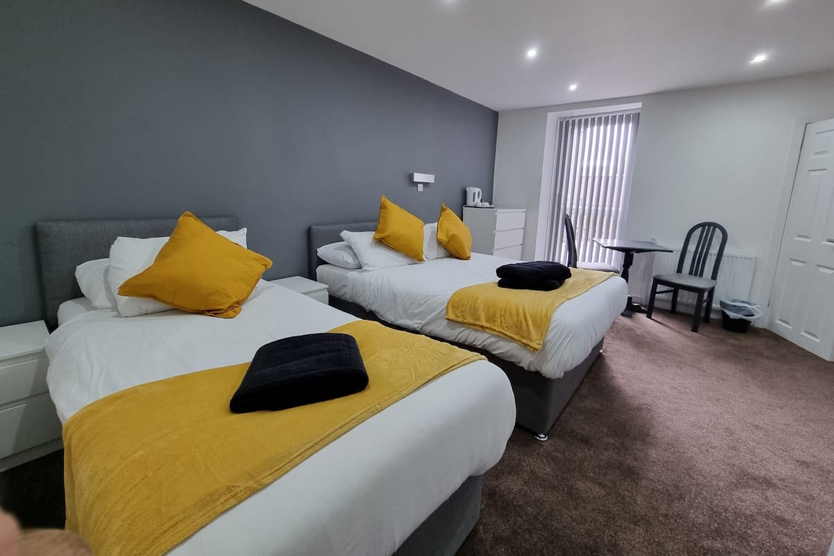 A hotel room with two beds, each with white bedding and yellow pillows, a small table, a chair, and a radiator.