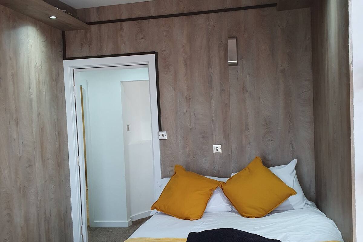 A modern hotel room with wooden panel walls, a bed with white bedding and two mustard pillows, a black towel, and a door leading to another room.