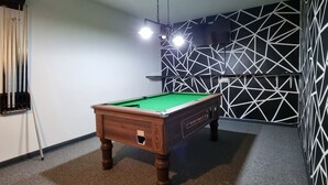 Ein Billardtisch in einem Raum mit geometrischem Wandmuster, einer Deckenleuchte und einem Teppichboden.