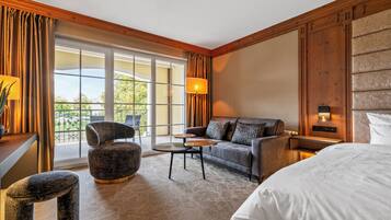 Ein Hotelzimmer mit einem großen Bett, einer Couch, einem Sessel, einem kleinen Tisch und einer Lampe. Ein Fenster mit Vorhängen bietet Blick auf Grünflächen.