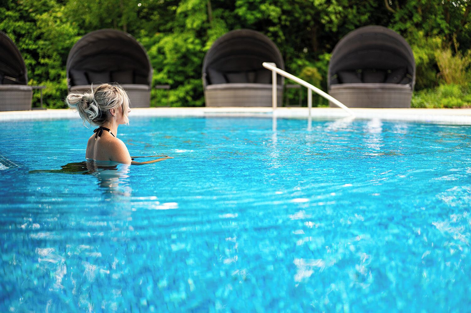 Eine Person in einem Swimmingpool mit klarem, blauem Wasser, umgeben von Grünpflanzen und befestigten Strukturen.