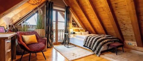 A cozy attic bedroom with a wooden ceiling, a plush red chair, a bed with a fur throw, and a large window with curtains.