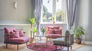 Una sala de estar acogedora con un sofá, un sillón y una mesita. Hay plantas en macetas y una vista al exterior a través de una ventana.