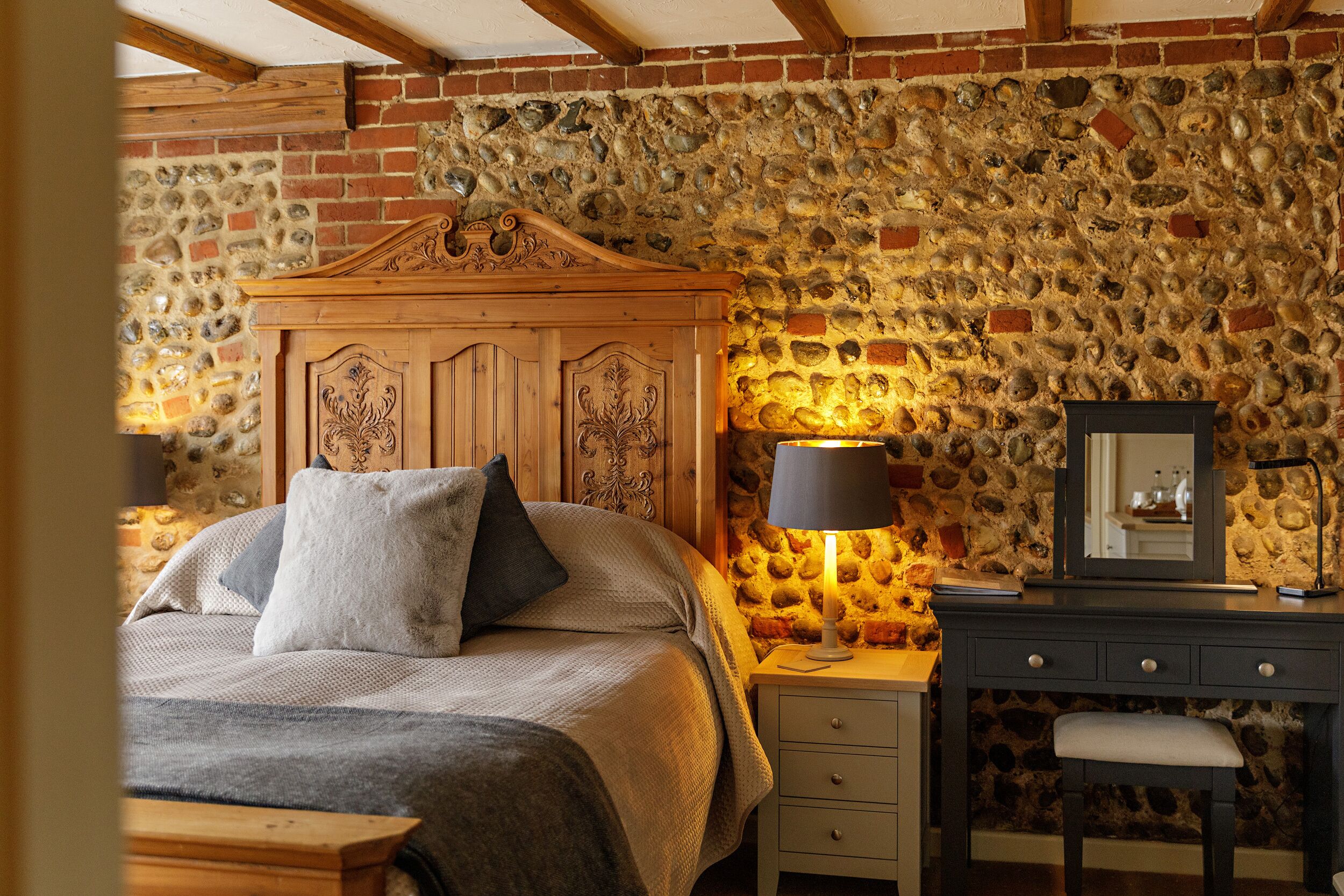 Un dormitorio con pared de piedra, cabecero de madera, mesita de noche con lámpara, espejo y una silla.