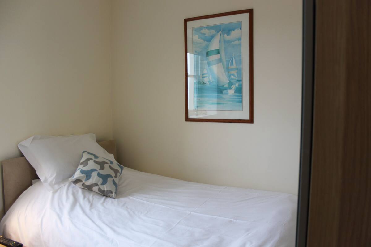 A neatly made bed with white linens and a single patterned pillow, a framed picture of sailboats on the wall, and a wooden door frame.