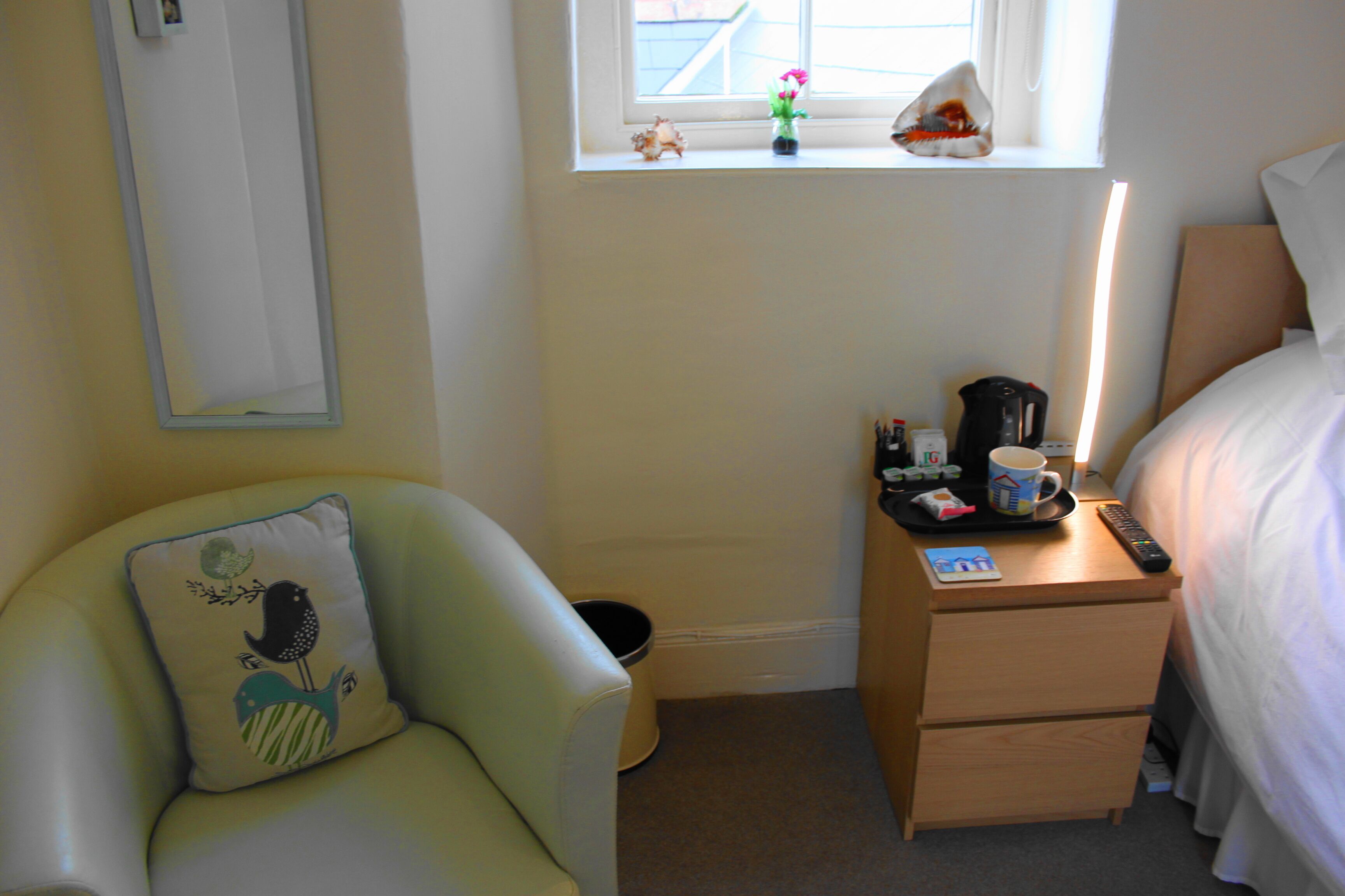 A hotel room with a bed, a nightstand, a chair with a pillow, a window with a view, and a small trash bin.