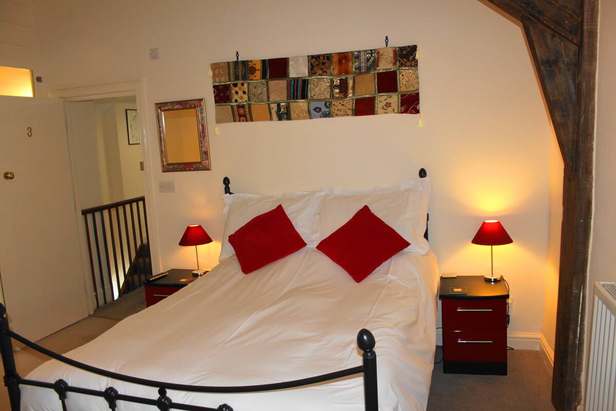 A neatly arranged bedroom with a bed, two bedside tables, a red lamp, a quilted tapestry, and a door with a number.