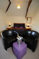 A room with a black leather sofa, a purple ottoman, a bedside table with a lamp, and a bed with purple bedding.