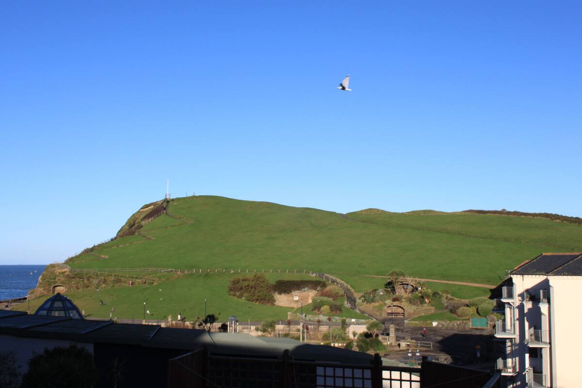 A coastal landscape with a hill, a building with a balcony, and a clear sky.
