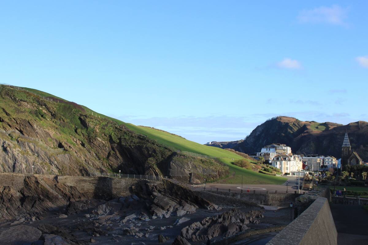 A coastal landscape with a rocky shoreline, a paved road, and buildings in the background.