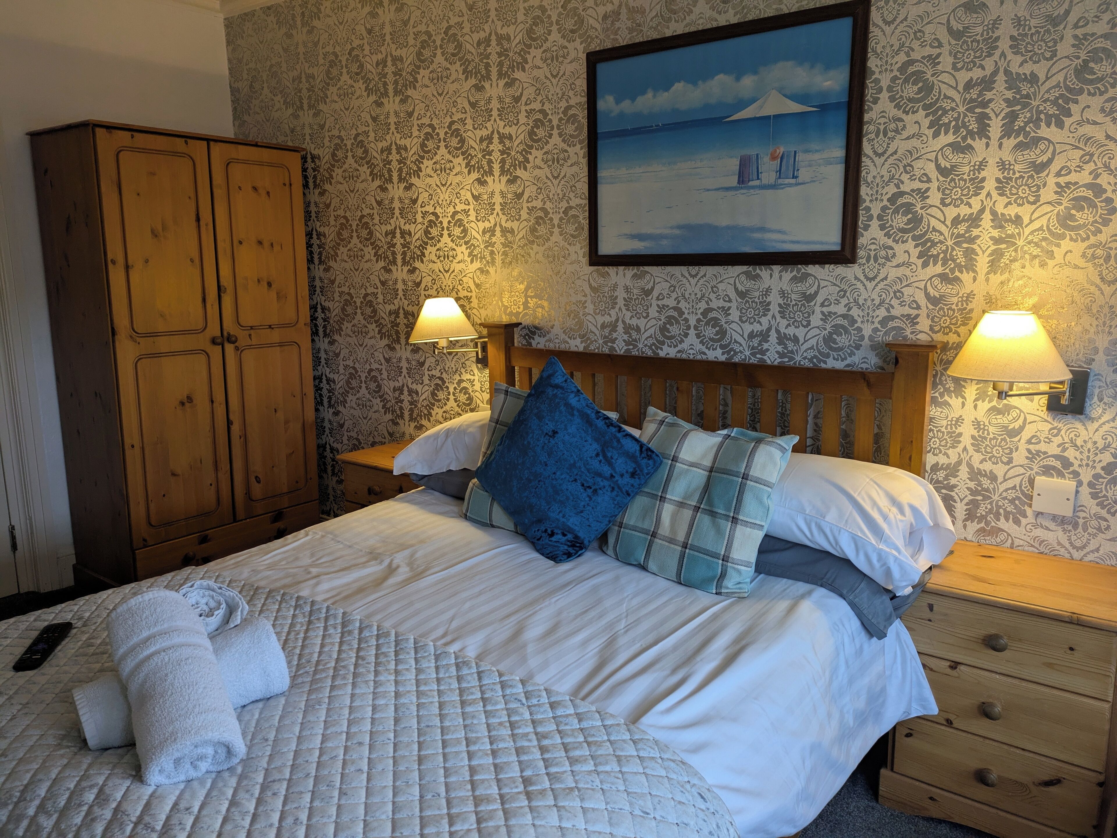 A neatly made bed with a quilt, pillows, and rolled towels. There is a wooden wardrobe and a painting of a beach scene on the wall.