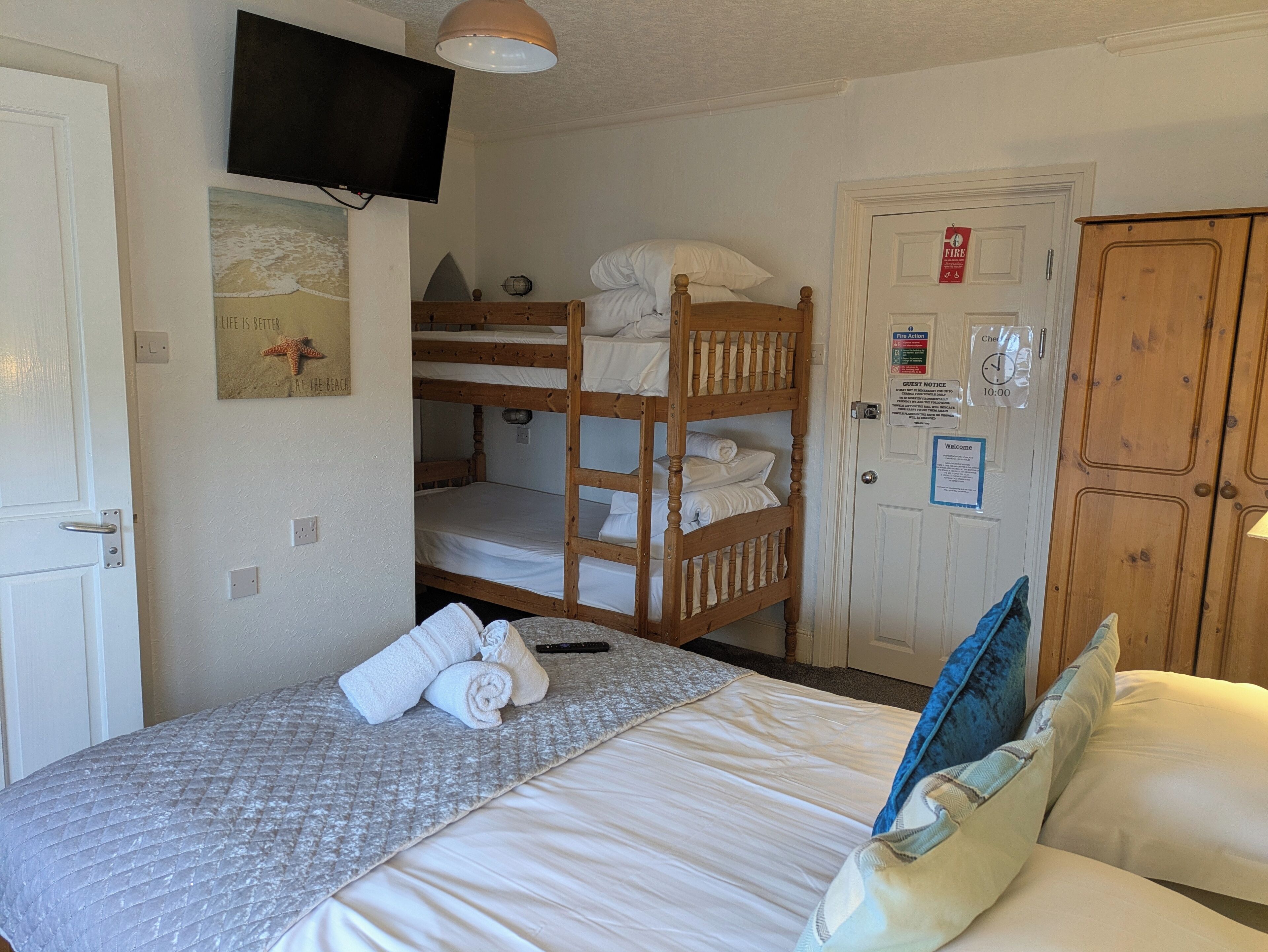 A room with a bunk bed, a single bed with a quilt, a flat-screen TV, a clock, and a door with a fire action sign.